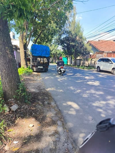 The image shows a street in Baleendah, Indonesia, with coordinates -7.006 latitude and 107.628 longitude. A large tree stands on the left side of the street, its shadow cast on the ground. A blue tarp-covered truck is parked on the right side of the street. A man on a motorcycle rides past the truck in the direction of the camera. A white car is parked on the right side of the street, behind the truck. The street is paved with asphalt and appears to be empty of pedestrians. The surrounding area is residential with houses and buildings visible in the background. There are many overhead power lines.  The sky is blue and clear.