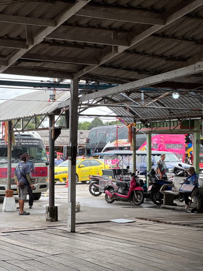 The image depicts a covered outdoor market or transportation hub in Ban Phe, Thailand. The foreground features weathered wooden planks, suggesting a pier or boardwalk. In the midground, various vehicles are visible, including a white bus with Thai text, a bright yellow taxi, a pink bus, and a blue SUV. Several people are present: two men are standing near the white bus, one of whom appears to be a driver or attendant; another man is standing near the yellow taxi; and a woman is seated on a bench, looking down at something in her lap. Multiple motorcycles and scooters are parked throughout the scene. Overhead, a corrugated metal roof supported by metal beams creates a shaded area. The background shows more buildings and trees, with an overcast sky. The overall impression is a bustling, utilitarian public space.