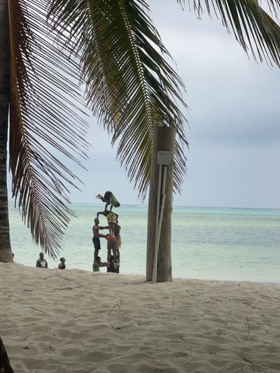 A daytime photograph captures a beach scene in Kiwengwa, Tanzania, under an overcast sky. The lower half of the frame displays light-colored sand with scattered small organic debris. Two palm tree trunks are visible on the left, with their fronds extending into the upper left and central top portions of the image. On the right, a weathered wooden utility pole stands erect, featuring a light-grey electrical box and two thin conduits attached to its side.
In the midground, at the immediate edge of the shallow, turquoise ocean water, approximately seven to eight males are engaged in forming a multi-tiered human pyramid. The base of the pyramid consists of at least three individuals partially submerged, supporting a second tier of two to three individuals. At the apex of the pyramid, one individual, wearing a dark green or black shirt and dark shorts, is performing an inverted acrobatic pose, possibly a handstand or pike, with legs bent. Two additional males are visible standing in the shallow water to the left of the human pyramid. The ocean extends into the background with a gradient from lighter turquoise near the shore to a deeper blue-green further out, meeting a uniformly light grey, overcast sky at the horizon.