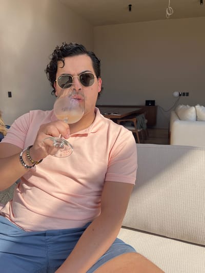 A man lies on a cushioned surface, wearing a pink polo shirt, blue shorts, and sunglasses. He holds a wine glass with a light-colored beverage. Bracelets adorn his wrist. The background includes a room with minimal decor, featuring a sofa, table, and visible wiring. The image is rotated.