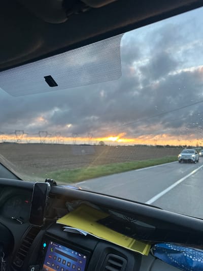 The image is taken from inside a vehicle, likely a car or truck, viewing the road ahead through the windshield. The sky is visible through the windshield, showing a sunset with a mix of clouds and clear areas where the sun's light shines through. Power lines are visible on the horizon, silhouetted against the setting sun. The road appears to be straight and flat, with a grassy shoulder on at least one side. Another vehicle is visible in the distance on the same road. Parts of the vehicle's dashboard are visible, including a digital display and some papers. The location is speculated to be Marcoussis, France, based on the OCR text.