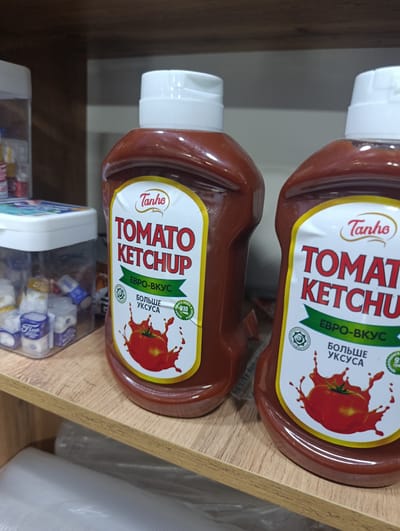 The image shows two bottles of Tanho Tomato Ketchup on a wooden shelf. The bottles are red with white caps and feature a label with a yellow border, red text, and an illustration of a tomato. The left bottle's label reads "TOMATO KETCHUP" in large red letters, with "Tanho" in smaller red script above it. Below, it says "ЕВРО-ВКУС" in green and "БОЛЬШЕ УКСУСА" in black. The right bottle's label is partially visible but appears to have similar text and design. To the left of the ketchup bottles, there is a clear plastic container filled with small, individually wrapped items, some of which are labeled "Five". The shelf is made of light-colored wood and is positioned against a dark wooden background. The scene appears to be a retail environment, possibly a shop or market stall, located in Khuroson, Tajikistan.