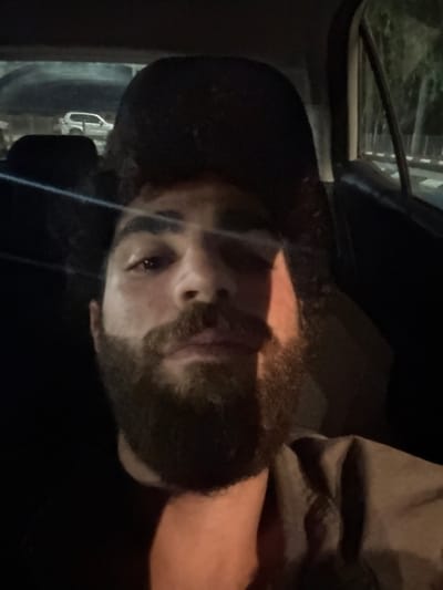 A male individual is depicted in a dimly lit vehicle interior, facing forward. He has dark, curly hair and a full, dark beard. A dark baseball cap is worn backward on his head. He is wearing a dark undershirt and a lighter, possibly olive-colored, collared shirt or jacket. A bright light source illuminates the left side of his face (viewer's perspective), casting a distinct shadow across the right side. A horizontal light streak is visible across his upper face. Through the rear window, a light-colored SUV is visible in the distance. Through the right-side window, trees and what appears to be an urban or suburban street at night are visible. The location is Brener Regional Council, Israel.