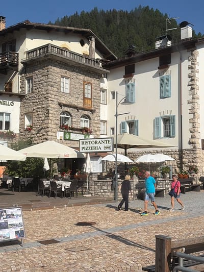 The image depicts a street scene in Campitello di Fassa, Italy, on a sunny day. A two-story building with a stone facade and a cream-colored upper story is prominent, featuring balconies, arched windows, and blue shutters. Adjacent to it is a white building with similar blue shutters and stone accents. In front of the buildings, an outdoor seating area for a restaurant is set up with tables covered in patterned cloths and chairs. Several large umbrellas provide shade. A sign indicates "RISTORANTE PIZZERIA."
Three people are walking across the cobblestone street. A woman in a black outfit walks away from the camera. A man in a blue polo shirt and denim shorts walks towards the right. A woman in a pink t-shirt and denim shorts follows behind him. In the background, a forested mountain slope rises under a clear blue sky. A display board with various images and text is visible on the left side of the foreground.