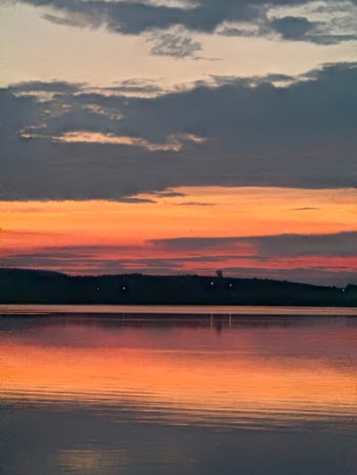This media file captures a serene sunset over a body of water. The sky is filled with dramatic clouds, painted in hues of deep gray, white, and vibrant orange and yellow as the sun dips below the horizon. The water reflects these colors, creating a stunning visual symphony of light and shadow.

Along the far bank of the water, a dark silhouette of trees and land is visible, with a few small lights suggesting distant human habitation or perhaps boats. A thin, straight line, likely a pier or a bridge, bisects the scene vertically, adding a geometric element to the organic landscape. The water's surface is rippled, distorting the reflections and adding texture to the scene.

The time of day is clearly sunset, indicated by the warm, low-angled light and the rich colors in the sky and water. The weather appears calm, with the gentle ripples on the water suggesting little to no wind. The overall emotion conveyed is one of peace and tranquility, a moment of natural beauty captured in time. No text is visible in the image.