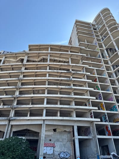 The image shows an unfinished, multi-story concrete structure in Perazića Do, Montenegro. The building's exterior consists of repeating horizontal slabs and vertical supports, creating numerous open floors and balconies. The raw concrete surfaces are heavily covered with various styles of graffiti, including tags, bubble letters, and more intricate designs in a range of colors. The sky is clear and blue. There is a dense green tree on the left side of the frame, partially obscuring the lower levels of the building. The scene suggests abandonment or a prolonged construction halt.