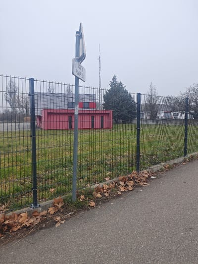 The image depicts an exterior scene in Hoenheim, France, under an overcast sky. In the foreground, an asphalt path or sidewalk is visible on the right, bordered by a strip of ground containing sparse green vegetation and numerous fallen brown leaves, particularly concentrated along the base of a black metal fence. This fence, composed of vertical bars and horizontal supports, extends across the midground, separating the paved area from an open grassy field.
A grey metallic pole is positioned in front of the fence, slightly to the left of the frame's center. Attached to this pole is a rectangular white sign with black text, oriented vertically, which reads "SORTIE POMPIERS" (Fire Exit / Firefighters' Exit).
Beyond the fence, the grassy field appears unmaintained, exhibiting variations in green and brown patches. In the background, a building complex is visible. The most prominent structure features a red lower section with multiple dark, rectangular window or panel openings, surmounted by an upper section with horizontal grey or silver siding and additional dark windows. To the right of this building, a large, dark green coniferous tree stands. Further in the distance, a tall, slender communication or transmission tower with a lattice structure is discernible against the light grey sky. Bare deciduous trees are also visible on both the left and right sides of the background.
No individuals, actions, or direct interactions are present in the scene. The overall lighting is diffuse due to the overcast conditions.