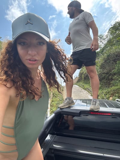 The image depicts a woman wearing a light-colored Jordan cap and a green swimsuit, likely a selfie, with a man standing on the open tailgate of a black SUV in the background. The man is wearing a light grey short-sleeved shirt and black shorts, and grey sneakers. He is standing on the tailgate with his back facing the camera. The surrounding environment appears to be a lush, green, possibly mountainous area. Power lines are also visible, and a glimpse of a paved road is visible behind the vehicle. Given the context, this is likely in a rural area of Suối Giàng, Vietnam.