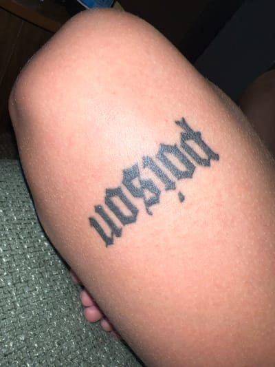 The image shows a person's right upper arm with a tattoo. The tattoo is of the word "wasgud" in a black gothic font. The arm is positioned on a dark gray fabric with a textured pattern. The image was taken in the city of Beersheba, Israel, at the coordinates 31.259, 34.778.  The image appears to be a close-up of a person's arm, with the subject's face and body not visible. The image is taken indoors, and the light is diffused and soft. The background is out of focus, with only a portion of the fabric visible.  The image quality is good.