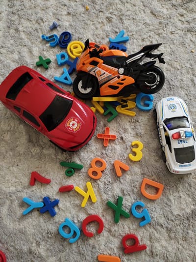 This media file showcases a collection of children's toys scattered across a textured, light-colored carpet. The primary objects include a red toy car with a "FIRE & RESCUE" emblem on its hood, an orange and black toy motorcycle with "HIGH SPEED" and "POLICE" decals, and a white toy police car with flashing blue and red lights and "PATROLMAN" written on its side.
Surrounding the toys are various plastic letters and numbers in multiple colors, including blue, yellow, orange, green, and red. Some of these include mathematical symbols like a plus sign and an equals sign, as well as letters such as "G", "D", "S", and "E". There are also several circular plastic items, one appearing to be an "@" symbol.
The arrangement suggests a play scene, with the toys and alphabet pieces seemingly placed or dropped on the carpet. The lighting appears to be overhead, creating a soft illumination across the scene, and there are no obvious indicators of time of day or weather conditions. The setting is a domestic indoor environment, likely a child's play area. No people are visible in the image. The visible text on the toys includes "FIRE & RESCUE", "HIGH SPEED", "POLICE", "PATROLMAN", and the number "7562".