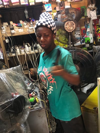 The image depicts an indoor scene in a cluttered electronics shop or charging station located in Enugwu-Ukwu, Nigeria. The central figure is a young adult, appearing female, with dark skin and a noticeable sheen on her forehead and face. She wears a black and white checkered beanie and a teal t-shirt with red and white text partially visible as "LIKE MONEY NO." Her right arm is blurred, indicating motion, with her hand appearing to manipulate a cable or connector.
Behind the individual, a wooden shelving unit occupies the left background, densely packed with various electrical outlets, multi-plugs, power adapters, and packaged electronic accessories. Numerous white and black cables hang vertically from these shelves, creating a tangled network. To the right of the central figure, a large black standing fan with a circular metal grill is prominently positioned, and another partial fan is visible further right. The right background also features shelves displaying packaged electronics.
In the foreground to the left, a counter or workbench holds items including a fan covered in clear plastic, a green object (possibly a multimeter), a yellow-handled screwdriver, and additional cables. The overall environment is functional, characterized by numerous electronic components and wiring, suggesting services related to charging or selling electronics.