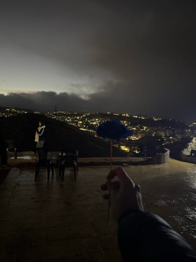 The image captures an elevated outdoor view of the city of Dummar Municipality, Syria, at dusk or night. The sky is overcast and dark, with hints of grey at the horizon. In the immediate foreground, a human hand holds a small, fluffy blue decorative object on a stick. The ground is a dark, tiled or paved surface, visibly wet and reflective. In the midground, to the left, a single person stands, mostly in silhouette, wearing a dark jacket and a light-colored head covering, facing away from the camera towards the city view. Two dark, plastic chairs are situated near this individual. A low concrete or stone wall separates the paved area from a dark, sloped hillside. The background displays a vast urban panorama, characterized by numerous illuminated buildings and streets spread across multiple hillsides, extending into the distant darkness under the cloudy sky.