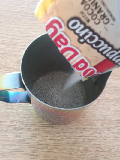 A pouch of instant cappuccino mix is being poured into a metallic pitcher on a wooden surface. The pouch, partially visible at the top of the frame, has red and beige lettering that reads "Cappuccino" and "Cocoa Gran" and "with". The pitcher is cylindrical with a handle that displays a rainbow of colors. The contents of the pitcher appear to be a dry, granular mixture. The lighting suggests an indoor setting, possibly a kitchen.