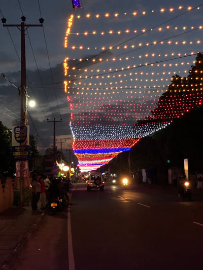 This media file depicts a festive street scene at dusk in Habaraduwa, Sri Lanka. The primary subject is a street adorned with a profusion of colorful lights, strung in elaborate patterns across the road and draped over trees. The lights range from warm yellow and orange to vibrant red, white, blue, and purple, creating a dazzling display against the darkening sky.
The scene includes several objects, such as utility poles with visible wires and insulators, and parked motorcycles. A few people are visible, some standing and observing the lights, while others appear to be socializing or moving along the street. The overall atmosphere suggests a celebration or a special event, possibly a festival or religious observance, given the elaborate lighting.
Notable details include the time of day, which appears to be twilight or early evening, with the sky transitioning from the muted tones of dusk to a darker hue. The weather seems clear. Location cues, like the Sri Lankan script on a sign, confirm the setting. The mood conveyed is festive and vibrant, with the lights adding a sense of joy and spectacle to the everyday streetscape.
Visible text includes Sri Lankan script on a sign attached to a utility pole. The text appears to be advertising something, with a phone number "0710 777 999" clearly visible. Another piece of text is also visible on a sign near the bottom right.