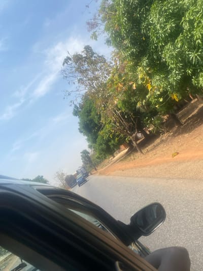 The scene depicts a roadside view in Bokana, Nigeria, observed from the interior of a dark-colored vehicle. The foreground, on the lower left, shows the window frame, upper door, and side-view mirror of the observer's vehicle. A partial view of a hand or arm is visible near the open window. The road, a grey asphalt surface, stretches into the distance, with a line of vehicles visible. These include at least two blue sedans and two large, dark-colored trucks, one bearing partially visible text "ALH MUK" on its side. To the right of the road, the ground is reddish-brown dirt. Further right and in the background, a dense line of trees with green and some browning foliage lines the roadside. The sky above is a clear, light blue with scattered wispy white clouds. The overall lighting suggests bright daylight conditions.