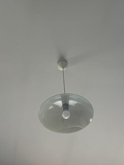 The image shows a close-up of a white ceiling with a single, modern pendant light fixture hanging from it. The fixture has a white circular canopy attached to the ceiling, from which a thin grey cord descends. At the end of the cord is a white socket holding a clear, round light bulb. The light bulb is enclosed by a large, translucent, dish-shaped shade made of glass. The shade has a delicate pattern of white, wavy lines etched or printed onto its surface. The background is the plain, textured white ceiling, with a soft shadow cast by the light fixture on the upper right side. The scene appears to be an interior setting, with no indication of the city of Marten, Bulgaria.