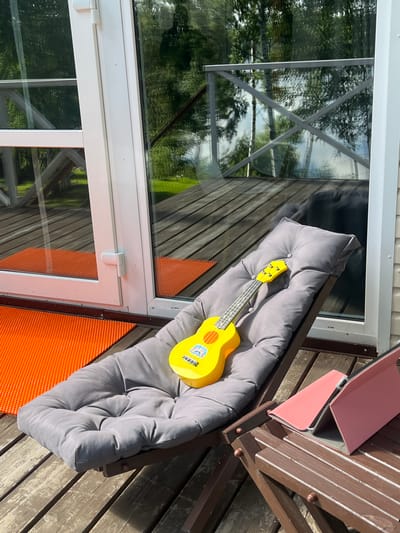 A yellow toy ukulele rests on a gray cushion on a wooden deck. Behind the deck is a glass door and railing with trees and a green lawn visible through the glass. To the right of the deck, a pink tablet case is open. The scene appears to be an outdoor setting in Naberezhnaya, Russia, likely a residential area.