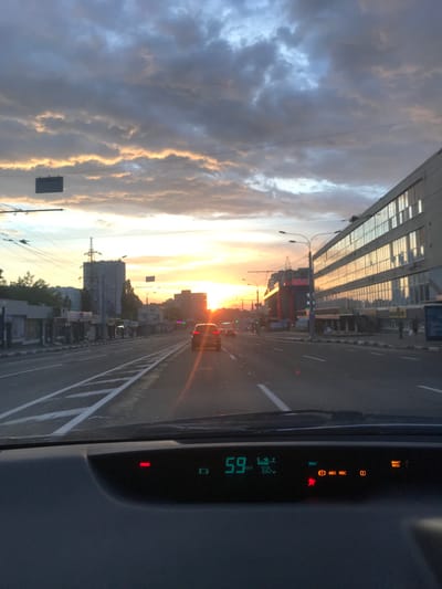 The image shows a car's dashboard displaying a speed of 59 km/h. The instrument panel illuminates several warning lights, including "BRAKE," "READY," "ABS VSC," "(!)," and "MAINT REQD." The view from the car's windshield reveals a cityscape at sunset. A road with lane markings stretches into the distance, with buildings lining either side. The location appears to be in the city of Babai, Ukraine. There's a car in front of the driver. The sky is filled with clouds illuminated by the setting sun.