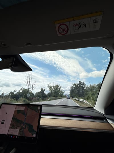 The image is taken from inside a vehicle, looking forward through the windshield. The sky is blue with scattered white clouds. The road ahead is paved and appears to be in a rural or semi-rural area, with trees and vegetation on either side. A van is visible in the distance, traveling in the same direction. The vehicle's dashboard is visible in the foreground, showing a large screen displaying a map and vehicle information. The car appears to be a Tesla, indicated by the design of the dashboard and screen. The location is identified as Ivanski, Bulgaria. There are no people clearly visible within the frame of the image. The scene depicts a typical view from a car driving on a road.