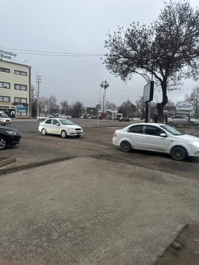 The image depicts an urban intersection in the city of Yangiiul, Uzbekistan, under an overcast sky. In the foreground, a rough asphalt and concrete surface covers the ground. Three cars are prominently featured: a black sedan partially visible on the far left, a white Chevrolet Aveo sedan positioned centrally and facing left, and another white sedan, blurred from motion, on the right, facing right. Multiple other vehicles, mostly white, are visible in the mid-ground and background, indicating active traffic.

To the left, a two-story commercial building displays "Hamkor Bank" signage on its upper facade and a lower sign with "BAXTLI OILAM TEAM" featuring wrestling figures. In the mid-ground right, a large, leafless deciduous tree stands next to a dark, rectangular billboard. Further in the background, a distinctive tall, white monument with a spherical top element is visible. Utility poles, overhead wires, and various bare trees populate the distant background, contributing to an overall winter or early spring aesthetic. No individuals are distinctly visible or engaged in direct interaction.