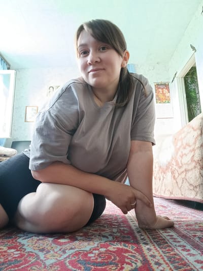 The image shows a woman in a room in what appears to be an apartment in Каменное, Russia. She is sitting on the floor, which is covered with a red and blue patterned rug. She is wearing a grey t-shirt and black shorts. Her hair is brown and pulled back in a ponytail. She is looking at the camera and smiling. In the background, there is a window with a view of trees. There is also a poster on the wall depicting two wolves. The room appears to be lit by natural light coming from the window. The overall impression is one of a casual, indoor setting.