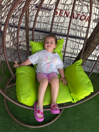 In Rusokastro, Bulgaria, a young girl sits in a hanging rattan chair filled with bright green cushions. She wears a grey t-shirt with colorful lettering that reads "JUST DO" and pink shorts. Her feet are adorned with pink sandals featuring strawberry designs. The chair is suspended outdoors, with a glimpse of what appears to be a boat with the word "SENATOR" visible in red lettering behind the girl. The ground beneath the chair is covered in artificial green turf.