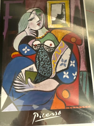 This media file is a close-up photograph of a framed print of a Cubist painting by Pablo Picasso. The painting depicts two figures, likely women, in a somewhat abstract and fragmented style characteristic of Cubism. One figure is lying down, with a pale face and green hair, resting her head on her arm. The other figure appears to be a seated woman wearing a blue garment and a patterned head covering. There are distinct blocks of color and geometric shapes throughout the artwork.
Notable details include:
* **Subjects:** Two female figures, a framed picture within the painting (yellow frame), and elements that suggest a domestic interior setting (though highly stylized).
* **Activity:** The figures appear to be in a state of rest or contemplation, with no active event clearly depicted.
* **Notable details:** The painting features bold colors like blue, red, orange, yellow, green, and purple. There are also floral motifs (white flowers on a blue background) and a patterned area in black and blue. The lighting in the photograph appears to be artificial, casting some glare on the surface of the print.
* **Visible text:** The signature "Picasso" is visible in white script on the right side of the print, indicating the artist.
The photograph is taken from a slightly angled perspective, and the print itself is enclosed in a plastic sleeve, evident from the reflections and the serrated edge at the top. The overall impression is that of a reproduction of a famous artwork. The location context provided is Chociule, Poland.