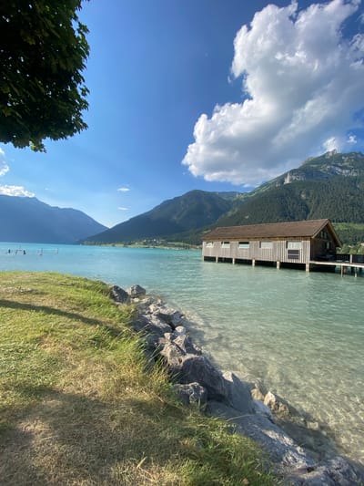 The image depicts a picturesque scene of a lake with a wooden structure built on stilts extending into the water. The structure, appearing to be a boathouse or a similar facility, has a pitched roof and multiple windows. The water is clear and a vibrant turquoise color, with sunlight reflecting off its surface. The shoreline is lined with rocks and lush green grass, with a few taller trees visible on the left side of the frame.

In the background, imposing mountains covered in dense green forests rise majestically. The sky is a brilliant blue, dotted with fluffy white clouds, suggesting a clear and sunny day. The overall atmosphere is serene and idyllic, evoking a sense of tranquility and natural beauty. The orientation of the image is landscape, with the horizon running vertically, suggesting it may have been captured with a phone that was held vertically.

The time of day appears to be daytime, likely late morning or afternoon, given the bright sunlight and clear skies. The location is identified as Lärchenwiese, Austria, which is known for its stunning alpine scenery and pristine lakes. There is no visible text in the image. No people are discernible in the frame, and there are no specific activities or events taking place beyond the serene display of nature.
