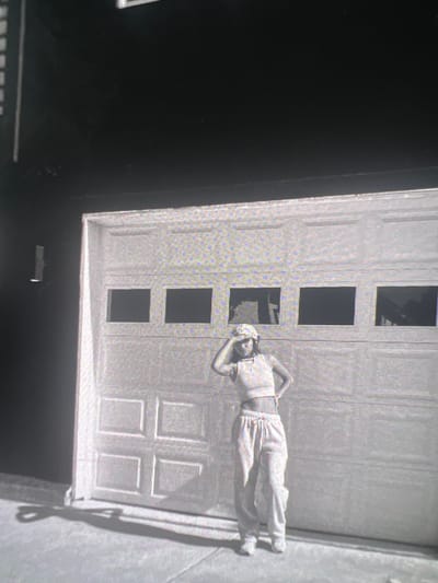 A black and white, full-body photograph shows a young woman standing in front of a closed garage door. The woman is wearing a cropped top, a bucket hat, and loose-fitting pants. She has her left hand on her forehead, shielding her eyes from the sun. The garage door is made of panels and has four windows across the top. A shadow of the woman is cast on the ground in front of her. The background is dark, suggesting it might be evening or a shaded area. The scene is set on a paved surface, possibly a driveway, in the city of Boone, United States of America.