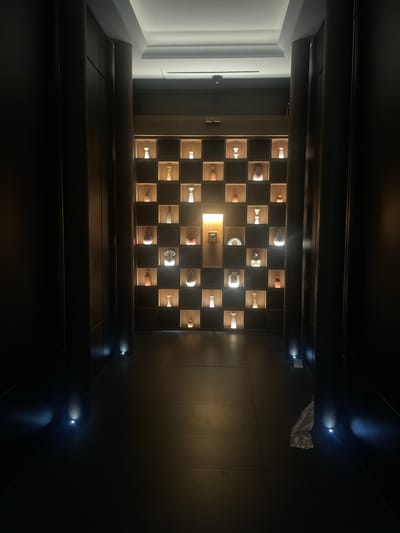 This is a wide-angle, low-light photograph taken inside what appears to be a modern, dimly lit hallway or passage, possibly in Gimpo-si, South Korea. The hallway is flanked by tall, dark wooden panels on both the left and right sides, which create a sense of depth and enclosure. The floor is tiled with large, dark square tiles.
The focal point of the image is a wall unit positioned at the end of the hallway. This unit is composed of a grid of individual cubbies, each illuminated from within. Within these cubbies are various decorative objects, predominantly vases and urns of different shapes, sizes, and colors, such as orange, white, and iridescent finishes. The lighting within the cubbies is warm and highlights the textures and forms of these displayed items. A bright, rectangular light source is centered behind some of the cubbies, creating a strong backlight effect.
Along the bottom of the hallway, embedded in the floor at intervals, are small, circular spotlights casting pools of blueish light onto the tiled floor, further accentuating the architectural lines and the polished surface.
In the far background, near the ceiling, a security camera is visible. On the right side of the image, near the floor, a crumpled piece of white material, possibly plastic sheeting or paper, is resting on the floor. There are no people visible in the photograph. The overall atmosphere is sophisticated and serene, with an emphasis on showcasing the displayed art objects through dramatic lighting.
