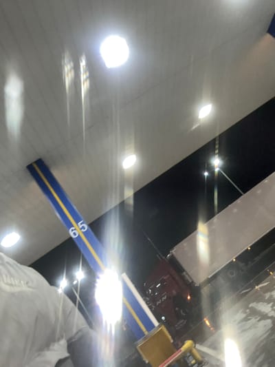 The image depicts an outdoor scene at night under an illuminated overhead canopy, identified as a location in Orizovo, Bulgaria. The canopy is light-colored, likely white or light gray, with multiple bright, circular recessed light fixtures casting strong illumination and reflections. A prominent structural support beam, colored blue with a yellow stripe down its center, displays the white numerical digits "65". Below the canopy, a dark red or maroon semi-truck cab with the letters "NN" visible on its side is parked, connected to a light-colored box trailer. The ground appears reflective, suggesting wetness. In the lower left quadrant, the upper back and shoulder of a person wearing a light-colored jacket are partially visible, facing away from the camera. Additional yellow and red equipment or structural elements are partially visible in the lower center foreground.