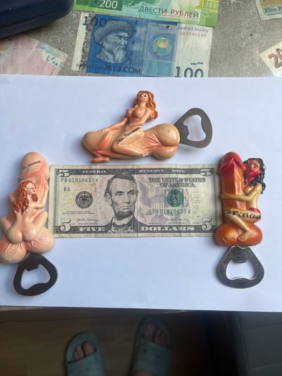 The image depicts a collection of novelty bottle openers with suggestive designs, laid out on a white surface, with several banknotes in the background. One bottle opener features a reclining female figure with "Sta. Susanna" inscribed on it. Another bottle opener shows a female figure embracing a phallic object, with "BARCELONA" written on it. A third bottle opener, partially visible, also appears to have a female figure and the inscription "Sta. Susanna". A US five-dollar bill is placed horizontally between two of the bottle openers, with Abraham Lincoln's portrait visible. In the background, partially obscured banknotes include denominations of 200 rubles and 100 Kyrgyzstani som, along with what appears to be a portion of a 20-unit banknote. The lighting suggests an indoor environment, potentially a tabletop or floor, with a dark, reflective surface visible at the bottom of the frame, possibly a piece of furniture. The context provided about Schwäbisch Hall, Germany, is not visually represented in the image itself.