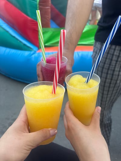 In Wershofen, Germany, an outdoor scene features three individuals holding colorful frozen drinks. The beverages, served in clear plastic cups, are a vibrant yellow and a pinkish-red, topped with ice-shaved textures. Each drink is adorned with a striped straw: green and white, red and white, and blue and white. In the blurred background, an inflatable structure with sections of blue, red, and green suggests a recreational setting, possibly a bouncy castle common at outdoor events or festivals in Wershofen. The image captures a moment of shared enjoyment, with the drinks and the playful background hinting at a festive atmosphere.