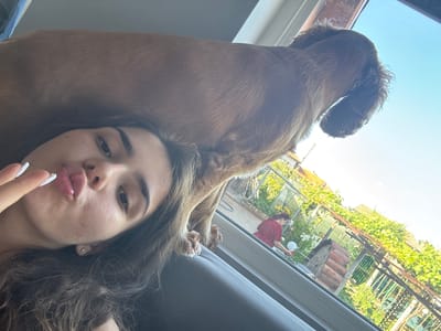 A low-angle, selfie-style shot captures a young woman with long, dark, wavy hair lying down, making a kissing gesture with her lips. Her right hand is raised near her face, with fingernails painted white. A brown dog is perched behind her head, its back obscuring the top of her head. The dog appears to be looking out of a window. Through the window, a sunlit garden is visible, with lush green foliage and some structures. A woman in a red shirt is seen in the garden, holding a long-handled tool, possibly watering plants. The scene is set against a bright blue sky. The location is identified as Dibich, Bulgaria.