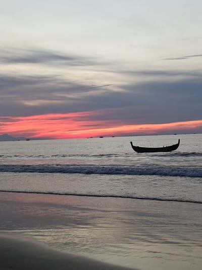 A long, narrow boat is silhouetted on the calm ocean water. It is facing right and appears to be empty. The sun is low on the horizon, casting a vibrant pink and orange glow across the sky. The water is a deep blue, with gentle waves lapping at the shore in the foreground. The beach is dark and reflective, mirroring the colors of the sky. In the distance, a faint outline of mountains can be seen. The scene appears to be tranquil and serene, capturing a moment of natural beauty at dusk. The location is identified as Curehtunong, Indonesia.