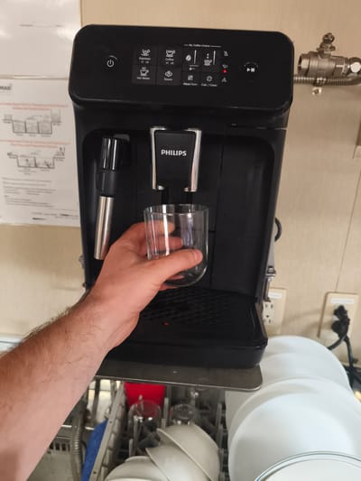 A person's arm and hand are visible, holding a clear glass under the spout of a black Philips brand automatic coffee machine. The coffee machine is positioned above a dishwasher, which is open and filled with dishes, including white plates and bowls. The scene appears to be indoors, likely in a kitchen or break room, with a beige wall and electrical outlets visible in the background. A printed diagram with instructions is mounted on the wall to the left of the coffee machine. The overall setting is functional and practical. The city of Velsen-Noord, Netherlands, is known for its industrial areas and port facilities. It is plausible that such a scene could be found in a canteen or common area within one of these facilities.