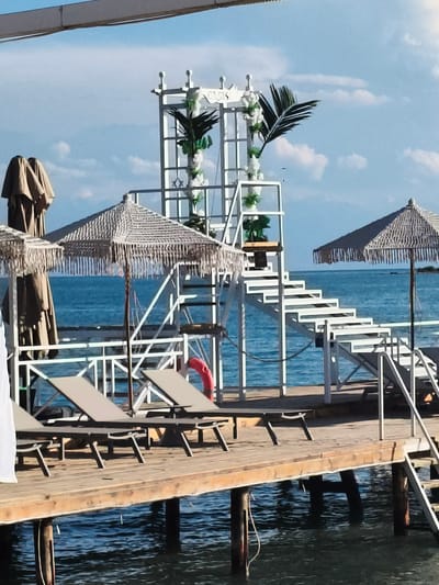 The image shows a wooden pier extending into a body of blue water, likely the sea or a large lake. The sky is blue with scattered white clouds. On the pier, there are several elements that suggest a resort or event setting.
In the foreground, there are two lounge chairs positioned on the wooden planks. One lounge chair on the left is partially visible, and another is more fully in view to its right. Behind the lounge chairs, there is a white structure with a white staircase leading upwards. This structure is decorated with greenery and white flowers, and it has a sign on top that is partially obscured but appears to read "GIS". A red life preserver ring is attached to the railing of this structure.
To the left of the central structure, there is a large, folded umbrella with a light brown fabric. To the right of the central structure, another similar umbrella is open, providing shade over a section of the pier. The water below the pier is calm and reflects the light. Wooden posts support the pier structure, and some are partially submerged in the water.
The overall scene evokes a peaceful and relaxing atmosphere, typical of a seaside vacation spot. There are no people visible in the image. The location in Bayet, Kyrgyzstan, is mentioned, though visually this scene is more consistent with a coastal region.