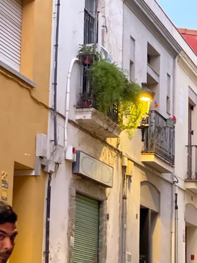 This image depicts a street scene in Santa Coloma de Gramenet, Spain, focusing on the facade of buildings. A man's face is partially visible on the left side of the frame, looking towards the camera. The buildings exhibit typical Mediterranean architecture with a mix of yellow and white stucco exteriors. On the white facade, there is a balcony adorned with lush green plants in a red planter, and an ornate street lamp emitting a warm yellow light. Below the balcony, a sign with faded lettering, possibly indicating a business, is mounted on the wall, and a green metal shutter is visible. Electrical conduits and junction boxes are present on the exterior walls, indicating the presence of utility infrastructure. The overall atmosphere suggests an urban environment, likely during the evening or late afternoon, given the artificial lighting.