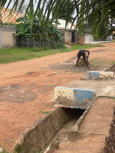 The image depicts a residential street scene in Kporva, Nigeria. A person, wearing black shorts with white stripes, is bent over on a reddish-brown dirt road, raking or gathering a pile of green leaves. The road appears to be in disrepair with patches of asphalt. To the right of the road, there is a concrete drainage ditch. Above the ditch are two rectangular concrete structures, one painted yellow and blue, the other primarily yellow, both showing signs of wear and peeling paint. In the background, there are houses with corrugated metal roofs and some vegetation, including banana trees. A barbed wire fence runs along the top of a wall on the left side of the frame. Palm fronds overhang the scene from the top.