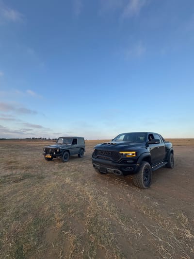 The image shows two vehicles parked on a dirt road in an open field. The sky is blue with some clouds. The field is covered in dry, light brown grass. The vehicle on the left is a gray Mercedes-Benz G-Wagon. The vehicle on the right is a black Ram 1500 TRX. Both vehicles are facing the camera. There are no people in the image. The coordinates provided are located near the city of Bnei Zion in Israel. The location is a rural area outside the city. The image appears to have been taken in the late afternoon.