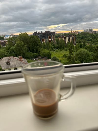 The image depicts a view from a windowsill, with a partially filled glass mug of coffee in the foreground, blurred due to shallow depth of field. Beyond the mug, the scene opens to a cityscape with a cloudy, overcast sky at dusk, casting a diffused light with hints of orange and yellow near the horizon. In the mid-ground, there are several multi-story apartment buildings with dark facades and numerous windows, interspersed with lush green trees and vegetation. A few older, low-rise buildings with grey, corrugated roofs are also visible, along with a red-tiled roof and chimneys. Further in the background, more apartment buildings and industrial structures can be observed. The overall impression is an urban or suburban setting, possibly during a transitional time of day. This scene is likely located in the city of Rubtsovo, Russia, given the context provided.