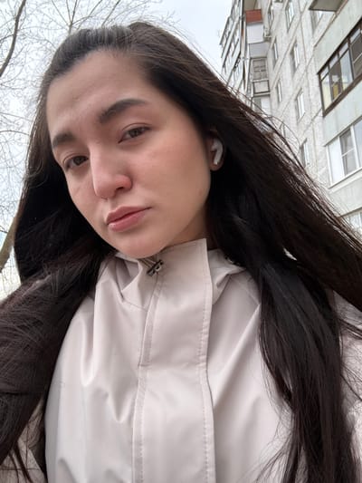 A selfie shows a young woman with dark hair and light skin wearing a white jacket. She has an earbud in her left ear. The background reveals a multi-story building with a repetitive window pattern, typical of Soviet-era architecture, suggesting the city of Kurovskoye, Russia. Bare branches are visible at the bottom of the frame.