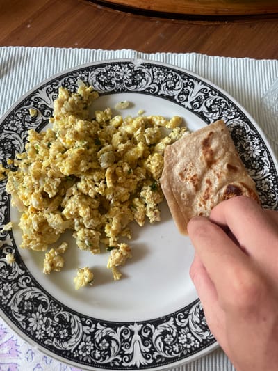 A top-down, medium shot shows a plate of scrambled eggs and a flatbread being held by a hand. The eggs, yellow in color, are piled on the left side of a white plate with an elaborate black floral border. A light brown, square-shaped flatbread is placed on top of the eggs. A hand is visible, holding one corner of the flatbread. The plate sits on a white striped placemat which is placed on a wooden surface. The location is likely a private residence or eatery in Chandigarh, India.