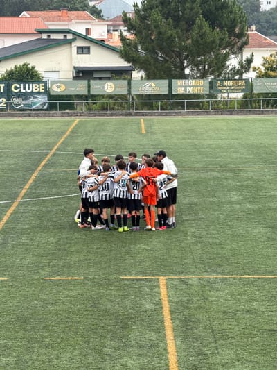 A group of young soccer players, wearing black and white striped uniforms and one in an orange goalkeeper uniform, are huddled together on a green turf soccer field. They are facing inwards, appearing to be in a huddle, possibly for a pep talk or a team discussion. A coach, wearing a white jacket and a black cap, stands within the huddle, with his arm around one of the players. The soccer field has yellow and white markings. In the background, there is a fence, banners with various advertisements and logos, trees, and buildings with red tile roofs. The scene appears to be outdoors, likely in a suburban or semi-urban area. The presence of Portuguese text on the banners suggests the location is in Portugal. Specifically, the banners mention "AJPRÍNCIPE MANUTENÇÃO E REPARAÇÃO DE AUTOMÓVEIS", "MERCADO DA PONTE", and "A. MOREIRA CONSTRUÇÃO - REMODELAÇÃO", all of which are common types of businesses found in Portuguese towns and cities. Given the specific businesses and the general architectural style, the image is likely located in Santa Maria da Feira, Portugal.
