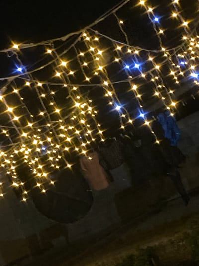 An outdoor scene at night is illuminated by numerous strings of warm yellow and some blue fairy lights, creating a bokeh effect against a dark background. The lights are draped and strung across the upper portion of the frame, casting a glow on objects and indistinct figures below. In the mid-ground, partially obscured by the lights, are several dark, silhouetted forms that appear to be people wearing dark clothing. One figure on the right is wearing a bright blue garment. To the left of center, a tan-colored object, possibly clothing, hangs amongst the lights. The ground appears to be dark pavement or dirt. The overall impression is that of a festive or decorated outdoor space at night. The location is the city of Babayurt, Russia.