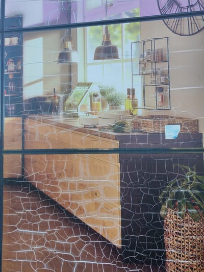 The image depicts a close-up view of a rustic kitchen scene, possibly a photograph or a mural, viewed through a cracked surface that distorts the image. The scene shows kitchen cabinetry, shelves with jars and bottles, hanging pendant lights, and a woven basket. The lower part of the image displays a surface with a cracked pattern that resembles aged wood or stone. There are no people visible in the image. The location information provided, Bielany Wrocławskie, Poland, is not visually verifiable from the image itself.