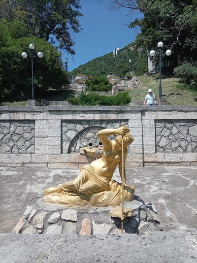 The image shows a golden statue of a reclining female figure, possibly a nymph or muse, in a park setting. The statue is seated on a rocky base and appears to be part of a fountain, with water spouts visible near its head. Behind the statue, there is a stone wall with decorative patterns and a long staircase leading up a hillside. Several lampposts with white globes are present along the path. In the background, a person in a white hat and blue shirt is visible walking up the stairs. The area is surrounded by lush green trees and vegetation, with a clear blue sky above. This scene is located in the city of Zheleznovodsk, Russia, known for its spa resorts and natural beauty.