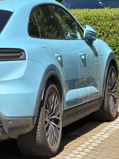 The image shows a light blue Porsche Taycan Cross Turismo parked on a cobblestone surface, likely in Fentange, Luxembourg. The focus is on the side of the car, highlighting its design elements like the door handles, side mirror, rear quarter panel vents, and the rear tire. The tires are Bridgestone. Green bushes are visible in the background, suggesting a residential or park area. There are no people visible in the image.