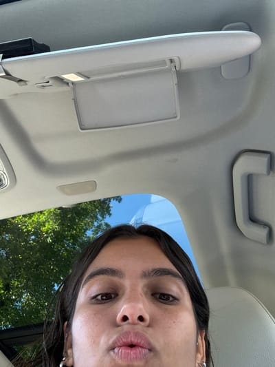 A close-up photograph, taken from inside a vehicle, captures a young person's face from the eyebrows down. The individual has dark hair and brown eyes, looking directly into the lens with pursed lips. They wear silver hoop earrings. The upper portion of the image shows the light-colored headliner of the vehicle, featuring a deployed sun visor with an illuminated vanity mirror on the driver's side and a light-colored grab handle on the passenger side. Through the car window, a clear blue sky and vibrant green tree foliage are visible. The specific location within Princeton, United States, cannot be determined from the visual information provided.