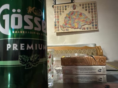 The image captures an indoor still life scene in Páty, Hungary, focused on a collection of items on a reflective dark surface against a light-colored wall. A large, metallic green can, labeled "Gösser PREMIUM" in white text with a hop-leaf emblem, occupies the foreground on the left. To its right, a clear glass with a repeating red cherry pattern is visible. Further right, a stack of three cigarette packs is present, with the topmost pack partially open. The side of these packs displays text in Hungarian, "CSAK FELNŐTTEKNEK ÁRUSÍTHATÓ" (FOR ADULTS ONLY), and English, "MADE IN THE EU UNDER THE AUTHORITY OF BRITISH AMERICAN TOBACCO (BRAND) INC. U.S.A." Behind the glass and cigarette packs, a light-colored wooden decorative item with a repeating geometric pattern rests on the surface. On the wall in the background, a hanging map of Hungary, segmented into various colors representing regions, is visible, framed by numerous small heraldic crests. A thin yellow cable is also present on the dark surface, extending from the right side. No individuals or active interactions are depicted.